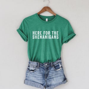 May include: A green t-shirt with the text "Here for the Shenanigans" in white lettering. The t-shirt is paired with denim shorts.
