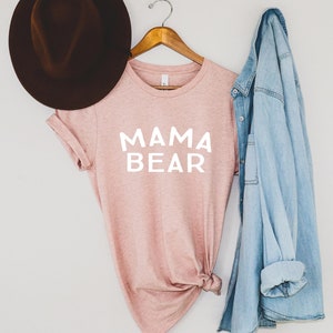 May include: A pink t-shirt with the text "MAMA BEAR" in white lettering. The shirt is hanging on a wooden hanger with a denim jacket draped over it. A brown fedora hat is on the left side of the image.