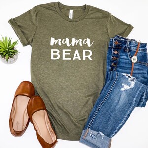 May include: A green t-shirt with the text "mama BEAR" in white lettering. The shirt is paired with blue jeans and brown flats.