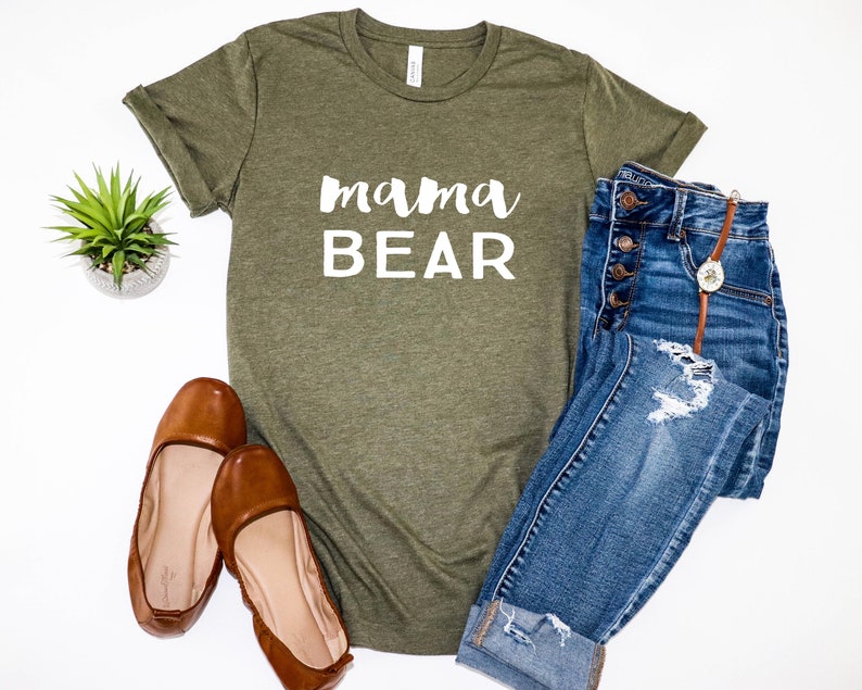 May include: A green t-shirt with the text "mama BEAR" in white lettering. The shirt is paired with blue jeans and brown flats.