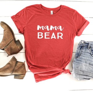 May include: Red t-shirt with the text "mama bear" in white lettering. The shirt is laid flat on a white wooden surface with a pair of brown ankle boots and a pair of blue denim shorts.