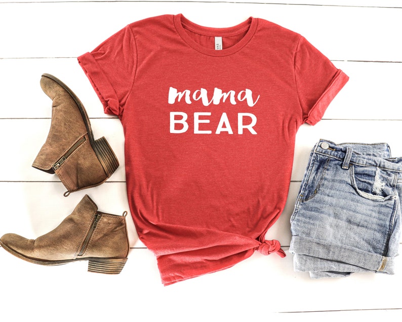 May include: Red t-shirt with the text "mama bear" in white lettering. The shirt is laid flat on a white wooden surface with a pair of brown ankle boots and a pair of blue denim shorts.