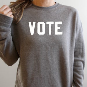 Peut inclure: Un sweat-shirt gris avec le mot "VOTE" imprim&eacute; en blanc sur le devant.