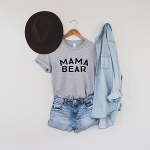 May include: A gray t-shirt with the text "MAMA BEAR" in black lettering. The shirt is paired with denim shorts and a light blue denim shirt. A brown fedora hat is on the left side of the image.