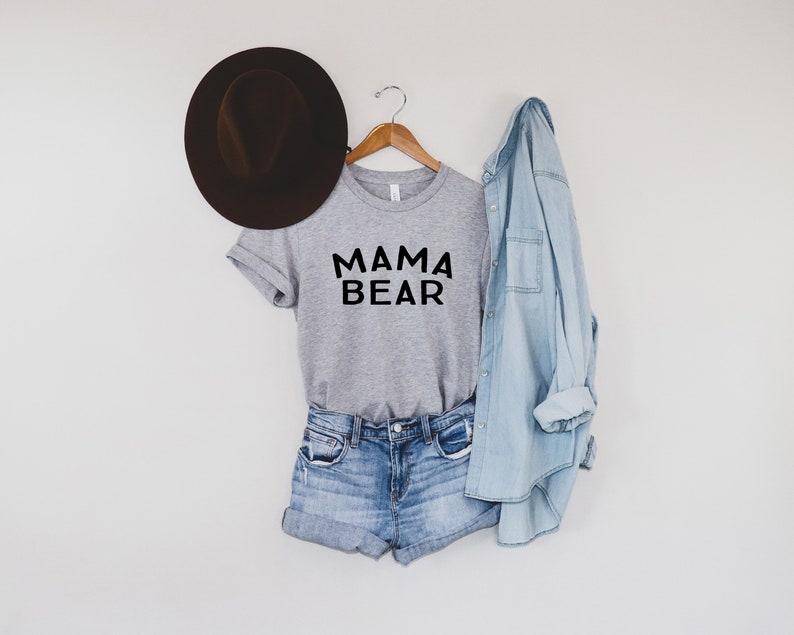 May include: A gray t-shirt with the text "MAMA BEAR" in black lettering. The shirt is paired with denim shorts and a light blue denim shirt. A brown fedora hat is on the left side of the image.