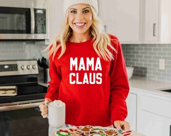 Op de afbeelding: Een vrouw draagt een rood sweatshirt met de tekst "MAMA CLAUS" in witte letters. Ze houdt een mok vast met een witte marshmallow erin en een bord met kerstkoekjes.