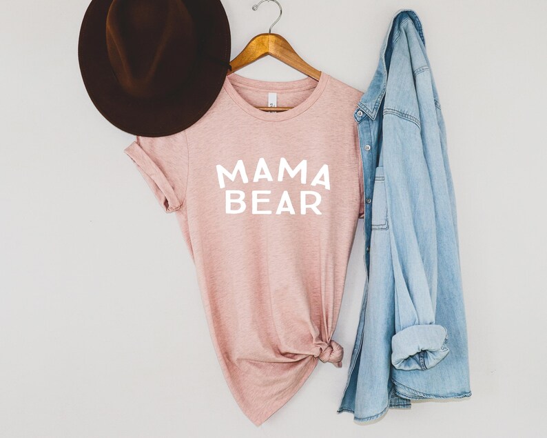 May include: A pink t-shirt with the text "MAMA BEAR" in white lettering. The shirt is hanging on a wooden hanger with a denim jacket draped over it. A brown fedora hat is on the left side of the image.