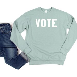 Peut inclure: Un sweat-shirt bleu clair avec le mot "VOTE" imprim&eacute; en blanc sur le devant. Le sweat-shirt est associ&eacute; &agrave; un jean bleu.