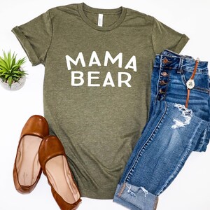 May include: A green t-shirt with the text "MAMA BEAR" in white lettering. The shirt is paired with blue jeans and brown flats.