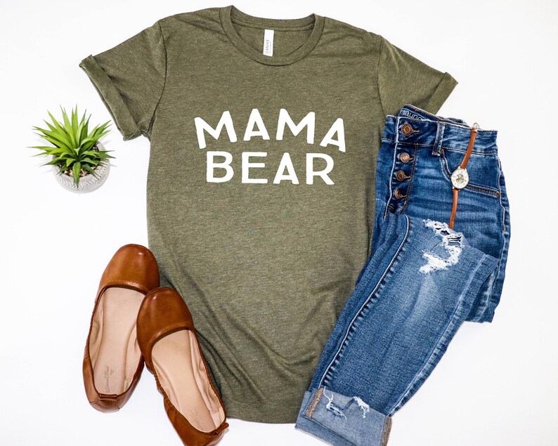 May include: A green t-shirt with the text "MAMA BEAR" in white lettering. The shirt is paired with blue jeans and brown flats.