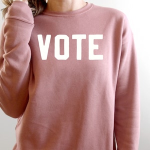 Peut inclure: Un sweat-shirt rose poussi&eacute;reux avec le mot "VOTE" imprim&eacute; en blanc sur le devant.