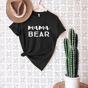 May include: Black t-shirt with white text that reads "mama BEAR" hanging on a hanger against a white wall with a black and white geometric pattern. A brown hat and a potted cactus are also in the image.