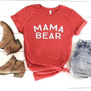 May include: Red t-shirt with the text "MAMA BEAR" in white lettering. The shirt is paired with denim shorts and brown ankle boots.