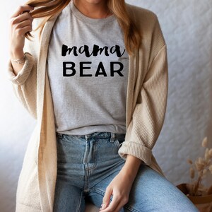 May include: A woman wearing a gray t-shirt with the text "mama BEAR" in black lettering. She is wearing a beige cardigan and blue jeans.