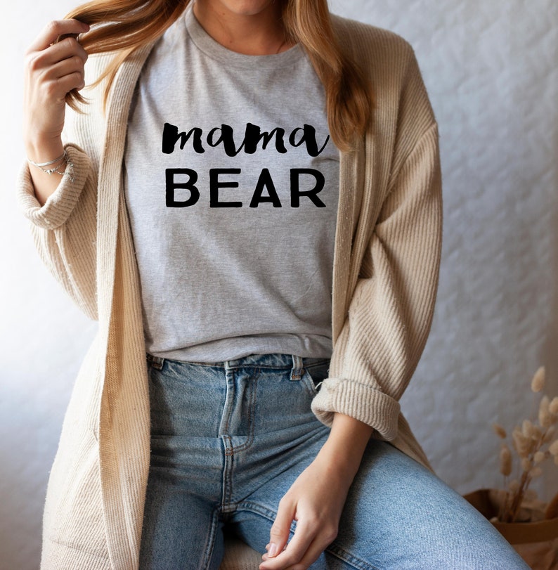 May include: A woman wearing a gray t-shirt with the text "mama BEAR" in black lettering. She is wearing a beige cardigan and blue jeans.