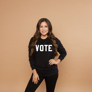 Peut inclure: Une femme portant un sweat-shirt noir avec le mot "VOTE" imprim&eacute; en blanc sur le devant. Elle est debout devant un fond brun clair.