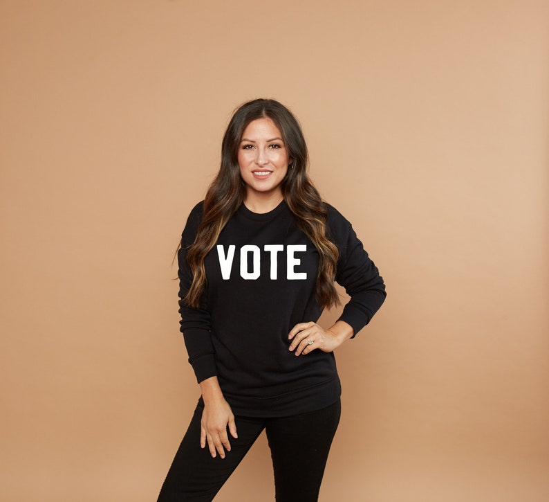 Peut inclure: Une femme portant un sweat-shirt noir avec le mot "VOTE" imprim&eacute; en blanc sur le devant. Elle est debout devant un fond brun clair.