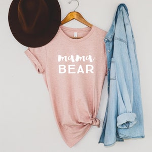 May include: A pink t-shirt with the text "mama bear" in white lettering. The shirt is hanging on a wooden hanger with a denim jacket and a brown hat.