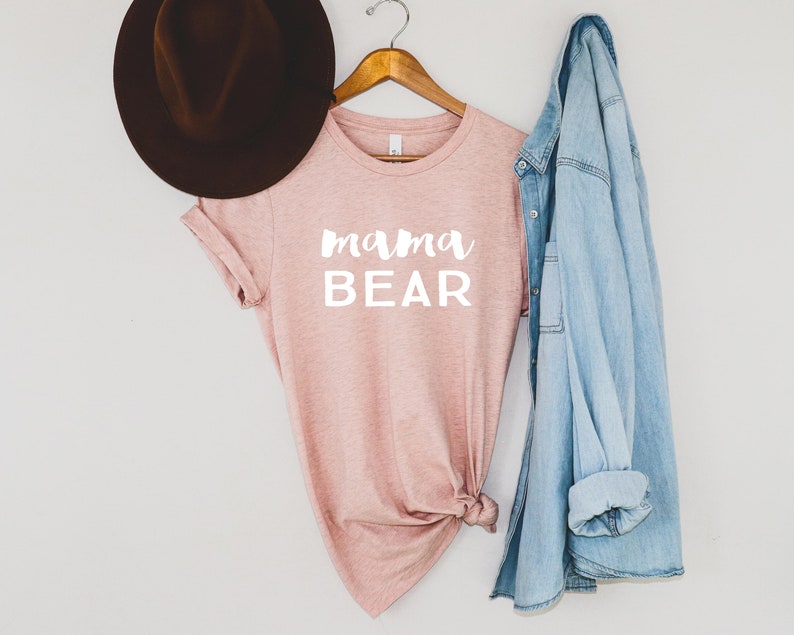 May include: A pink t-shirt with the text "mama bear" in white lettering. The shirt is hanging on a wooden hanger with a denim jacket and a brown hat.