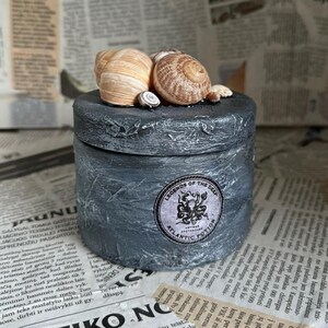 Può includere: Un barattolo di ceramica grigio con un coperchio nero e un adesivo bianco che dice "Legends of the Deep, Atlantic Potion". Il barattolo è decorato con conchiglie sulla parte superiore.
