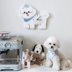 May include: A white ceramic wall clock shaped like a dog with blue and yellow accents. The clock is hanging on a white wall. There is a white dog sitting on a white table in the foreground.