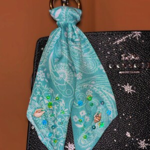 Puede incluir: Un pañuelo turquesa y blanco con estampado de cachemira y adornos de conchas y cuentas. El pañuelo está sujeto a un anillo metálico dorado. El pañuelo cuelga de un bolso negro.