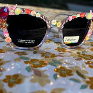 May include: Clear-framed sunglasses with black lenses, adorned with colorful embellishments. The frames are decorated with small charms, rhinestones, and the word "LOVE". The lenses have "ULTRAVIOLET PROTECTION" and "Polarized" labels.