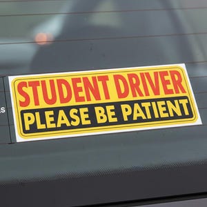 Op de afbeelding: Een rechthoekige gele sticker met zwarte en rode tekst: "STUDENT DRIVER PLEASE BE PATIENT". De sticker is ongeveer 10 cm hoog en is bevestigd op een autoruit.