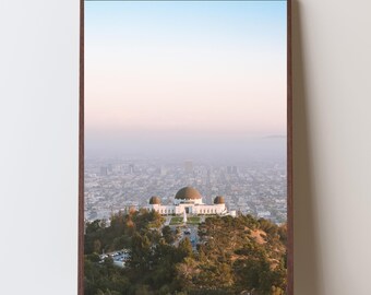 以下が含まれることがあります： ロサンゼルスのグリフィス天文台を写したフレーム入りプリント。丘の上に建つドーム型の屋根を持つ白い建物が写っており、背景には都市景観が、空には青とピンクのグラデーションが広がっています。フレームは濃い茶色です。