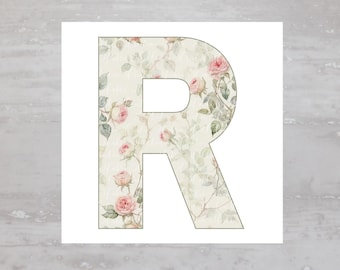 May include: The letter "R" featuring a floral design. The letter is filled with a pattern of pink roses, green leaves, and a cream-coloured background. The design is set against a white background.