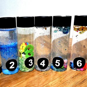 May include: Seven clear plastic sensory bottles with black screw-on caps, each filled with distinct liquid mixtures, colourful glitter, small plastic shapes, and numbered stickers from one to seven, arranged in a row on a wooden surface.