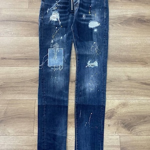 Może przedstawiać: Ciemne dżinsy z przetarciami, w tym podarte łatki i plamy farby. Posiadają zamek błyskawiczny z czerwoną zawieszką. Dżinsy mają dopasowany krój i są prezentowane na drewnianej powierzchni.