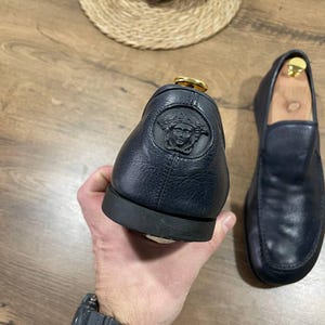 May include: Navy blue leather loafers, one held up to show a Medusa head emblem. The other shoe is on a wooden surface. The shoes have a gold-coloured accent.