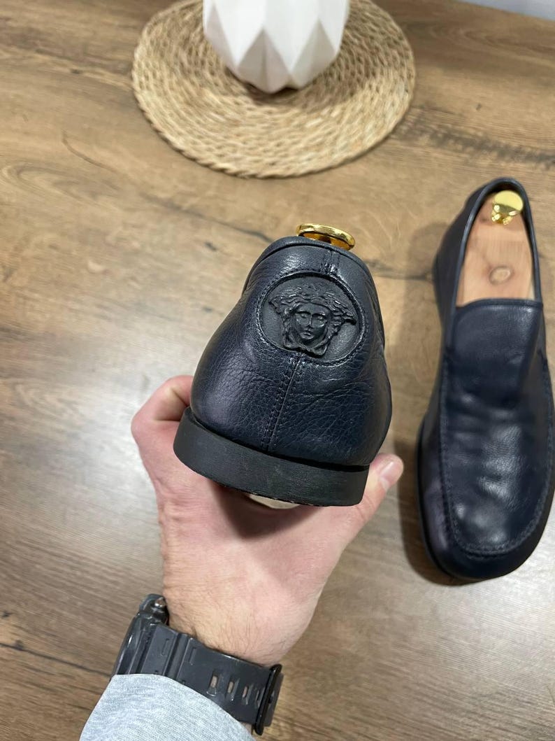 May include: Navy blue leather loafers, one held up to show a Medusa head emblem. The other shoe is on a wooden surface. The shoes have a gold-coloured accent.