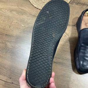 May include: Black sole of a loafer shoe with a textured, scale-like pattern. The shoe is held by a hand, revealing the underside of the shoe. The shoe is black and has a stitched edge.