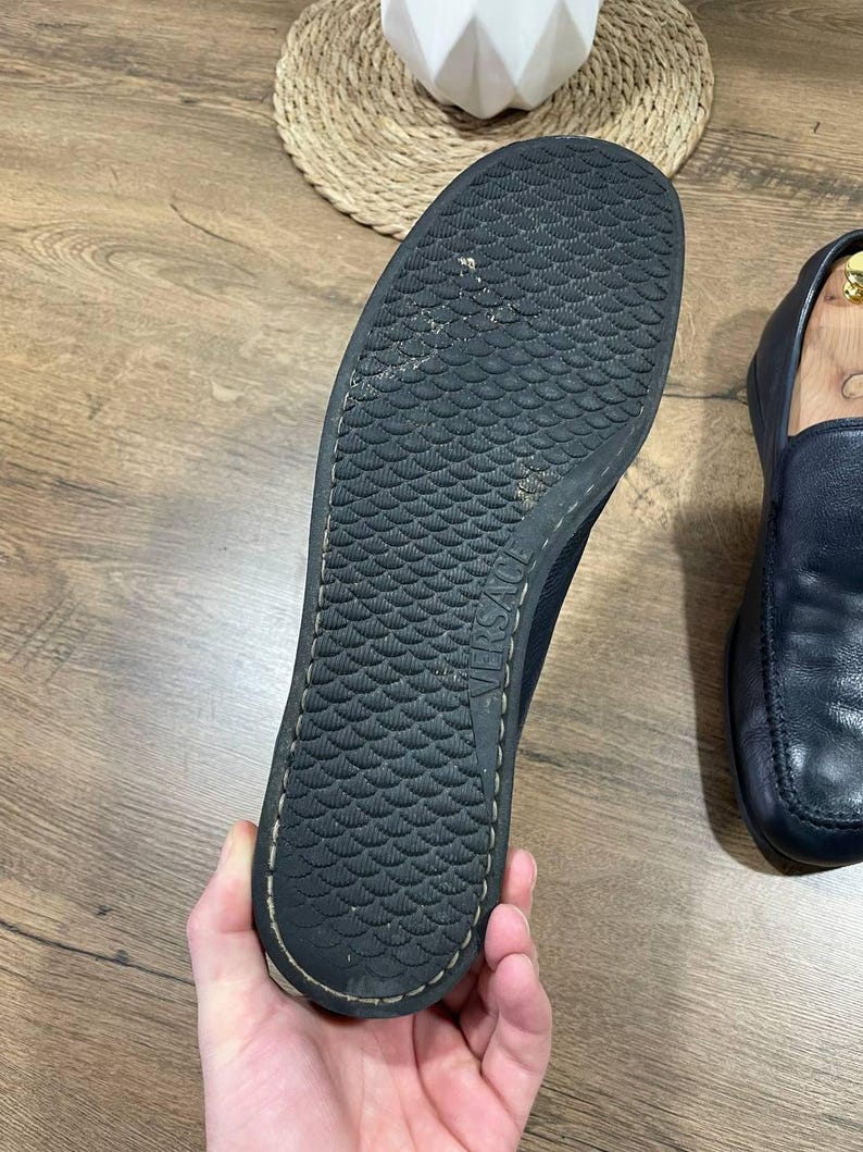 May include: Black sole of a loafer shoe with a textured, scale-like pattern. The shoe is held by a hand, revealing the underside of the shoe. The shoe is black and has a stitched edge.