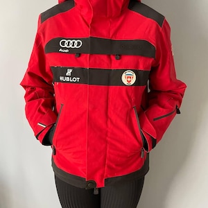 Puede incluir: Chaqueta de esquí roja con detalles y logotipos negros. La chaqueta presenta el logotipo de Audi, el logotipo de Hublot y un parche de la bandera suiza.