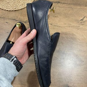 May include: Navy blue leather loafers with a rounded toe and a low, flat sole. The leather has a textured appearance. The shoes are displayed on a wooden surface.