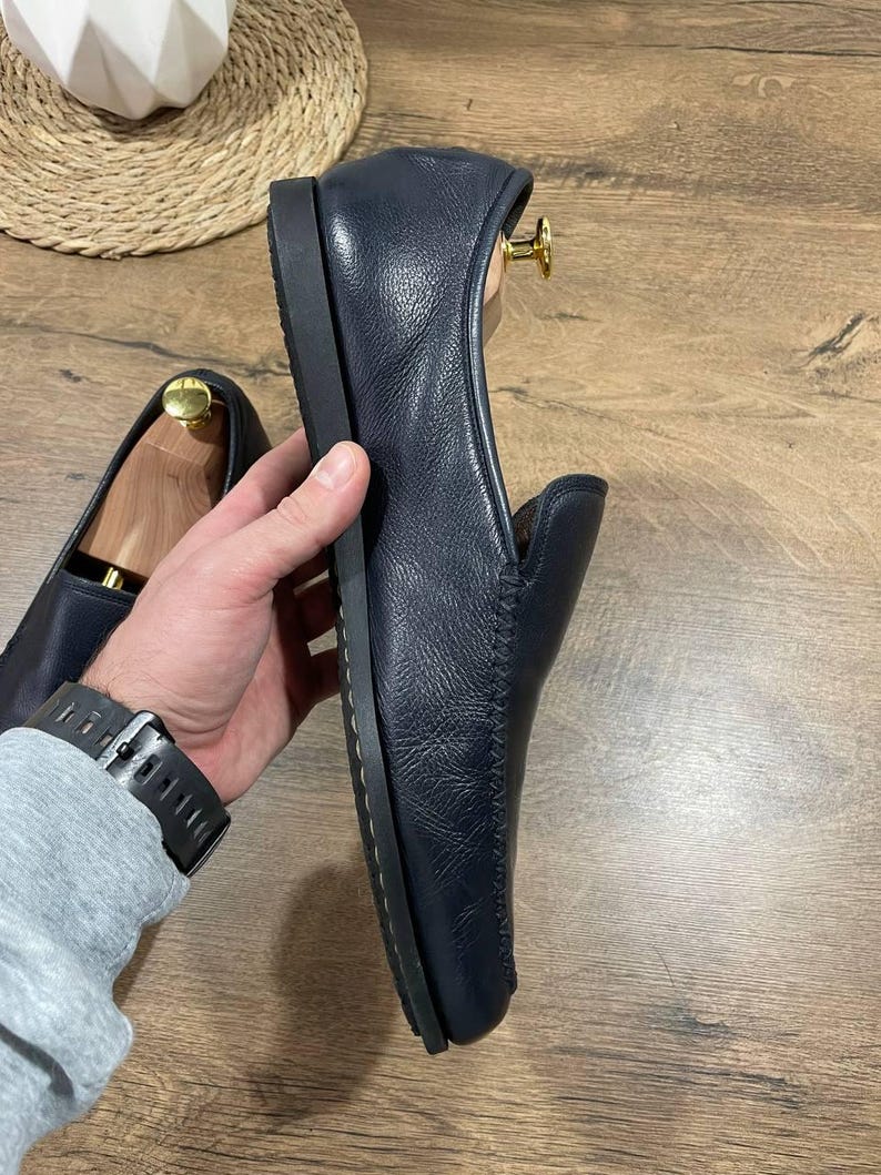 May include: Navy blue leather loafers with a rounded toe and a low, flat sole. The leather has a textured appearance. The shoes are displayed on a wooden surface.