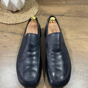 May include: A pair of black leather slip-on shoes with a rounded toe and stitched detailing. The shoes are displayed on a wooden surface, with wooden shoe trees inside. The shoes have a classic, minimalist design.
