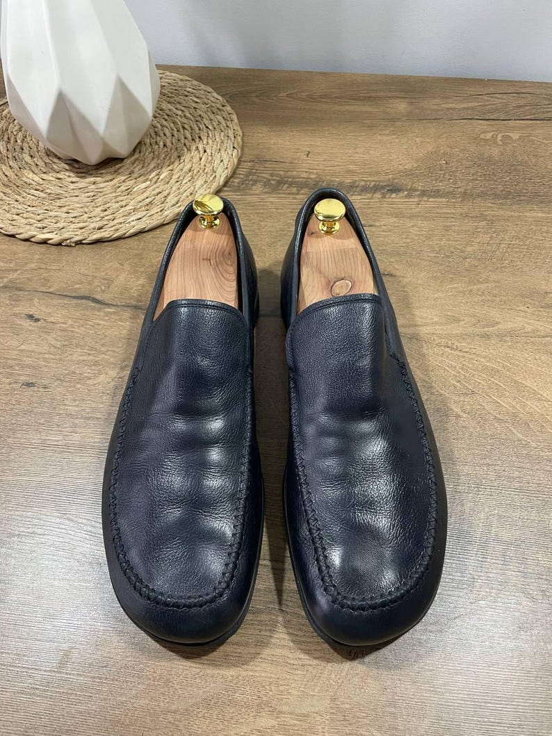 May include: A pair of black leather slip-on shoes with a rounded toe and stitched detailing. The shoes are displayed on a wooden surface, with wooden shoe trees inside. The shoes have a classic, minimalist design.