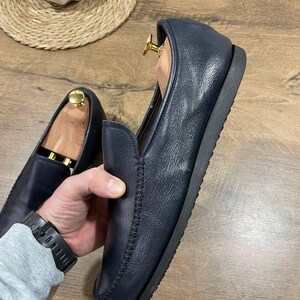 May include: Navy blue leather loafers with a stitched design. The shoes have a rounded toe and a black sole. Wooden shoe trees are inside the shoes. The shoes are on a wooden surface.