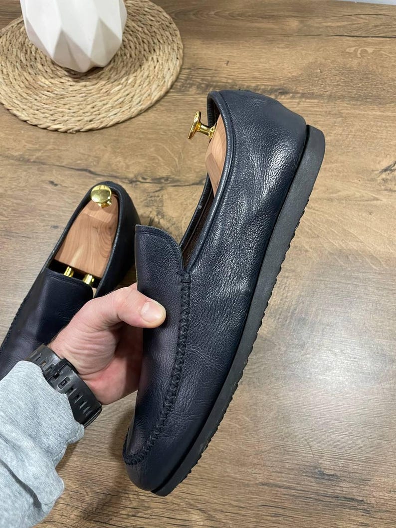 May include: Navy blue leather loafers with a stitched design. The shoes have a rounded toe and a black sole. Wooden shoe trees are inside the shoes. The shoes are on a wooden surface.