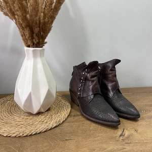 May include: A pair of dark brown leather ankle boots with a textured design on the toe and a folded-over top. The boots have a small heel and silver stud accents. A white geometric vase with dried plants is in the background.