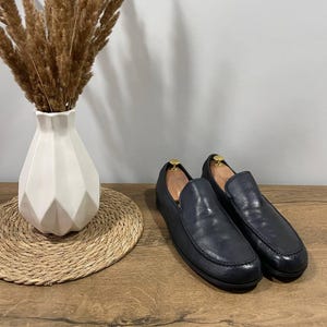 May include: Navy blue leather loafers with a classic design. The shoes are displayed on a wooden surface, next to a white geometric vase with dried ornamental grass and a woven placemat.