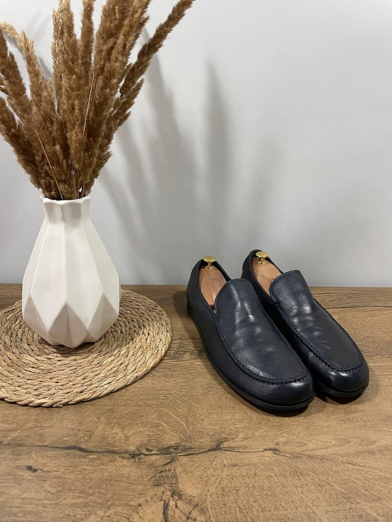 May include: Navy blue leather loafers with a classic design. The shoes are displayed on a wooden surface, next to a white geometric vase with dried ornamental grass and a woven placemat.