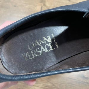 May include: Close-up of a dark blue leather shoe interior. The insole is brown with the gold text "GIANNI VERSACE" and "MADE IN ITALY". The shoe's edge is visible, showing the stitching and the quality of the leather.