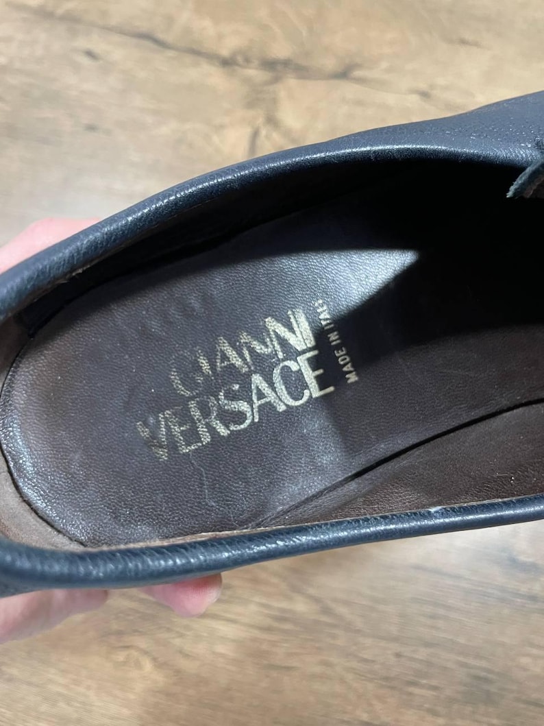 May include: Close-up of a dark blue leather shoe interior. The insole is brown with the gold text "GIANNI VERSACE" and "MADE IN ITALY". The shoe's edge is visible, showing the stitching and the quality of the leather.
