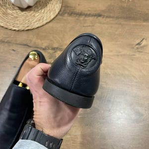 May include: Close-up of a black leather loafer with a Medusa head emblem on the heel. The shoe has a rounded toe and a black sole. The other shoe is visible in the background. The background is a wooden surface.