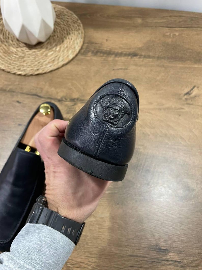 May include: Close-up of a black leather loafer with a Medusa head emblem on the heel. The shoe has a rounded toe and a black sole. The other shoe is visible in the background. The background is a wooden surface.
